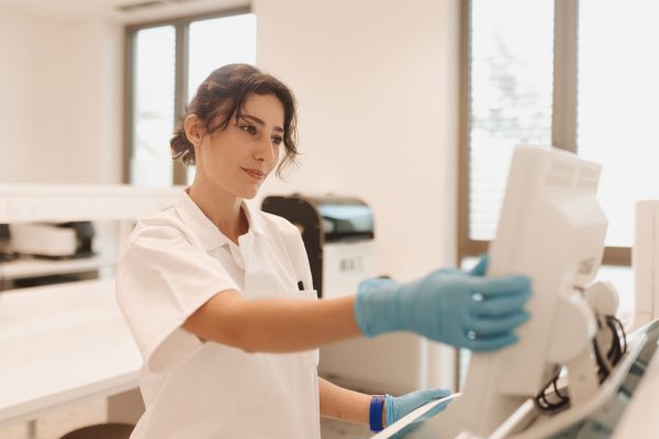 Žena u laboratorijskom okruženju nosi bijelu kutu i plave rukavice dok koristi računalo.