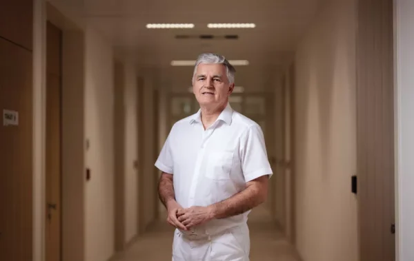 Stariji muškarac u bijeloj odjeći stoji u hodniku zgrade, osmijehujući se.