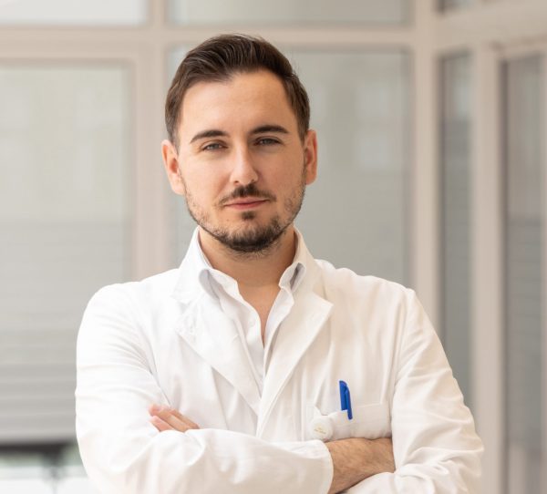 Muškarac u bijelom liječničkom ogrtaču s prekriženim rukama stoji u svijetloj prostoriji.