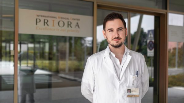 Muškarac u bijeloj liječničkoj kuti stoji ispred ulaza u medicinski centar s natpisom "PRIORA".