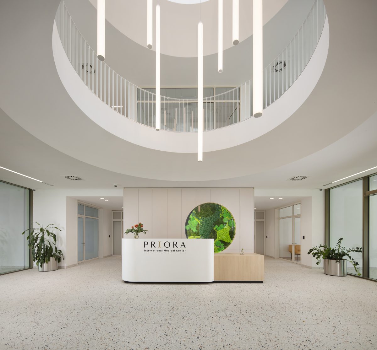Interijer modernog medicinskog centra s bijelom recepcijom koja nosi natpis 'PRIORA International Medical Center', okružen zelenim biljkama i osvijetljen visećim svjetiljkama.