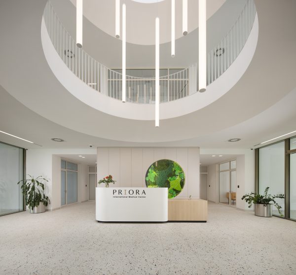 A modern reception area at a medical center with a curved white reception desk labeled "PRIORA International Medical Center," green plants, and numerous vertical hanging light fixtures under a circular ceiling opening.