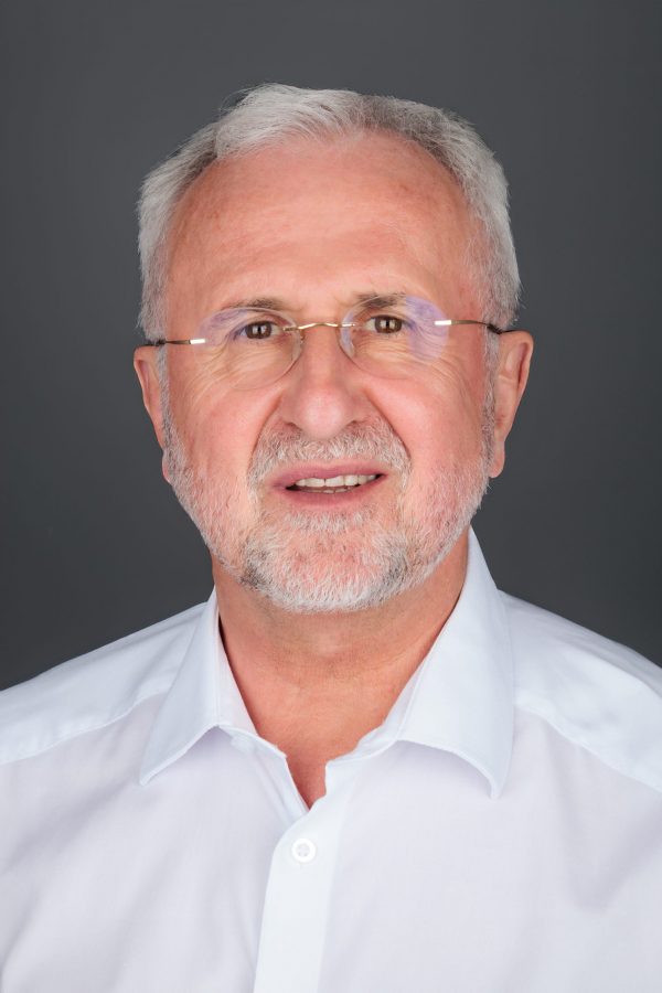 Stariji muškarac s bijelom kosom i naočalama nosi bijelu košulju, pozira ispred sive pozadine.