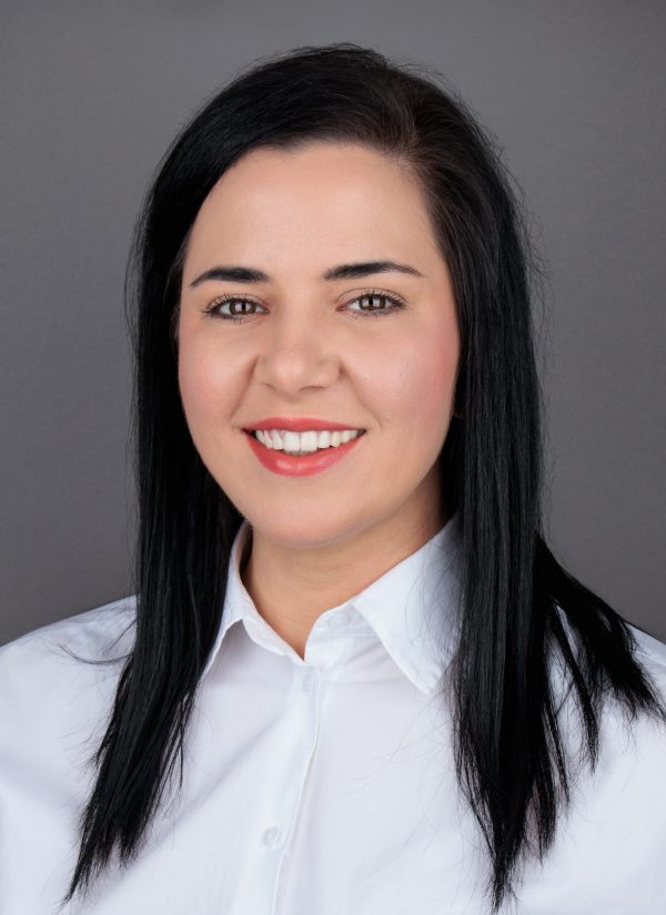 Žena s dugom crnom kosom nosi bijelu košulju i smiješi se, pozira pred sivom pozadinom.