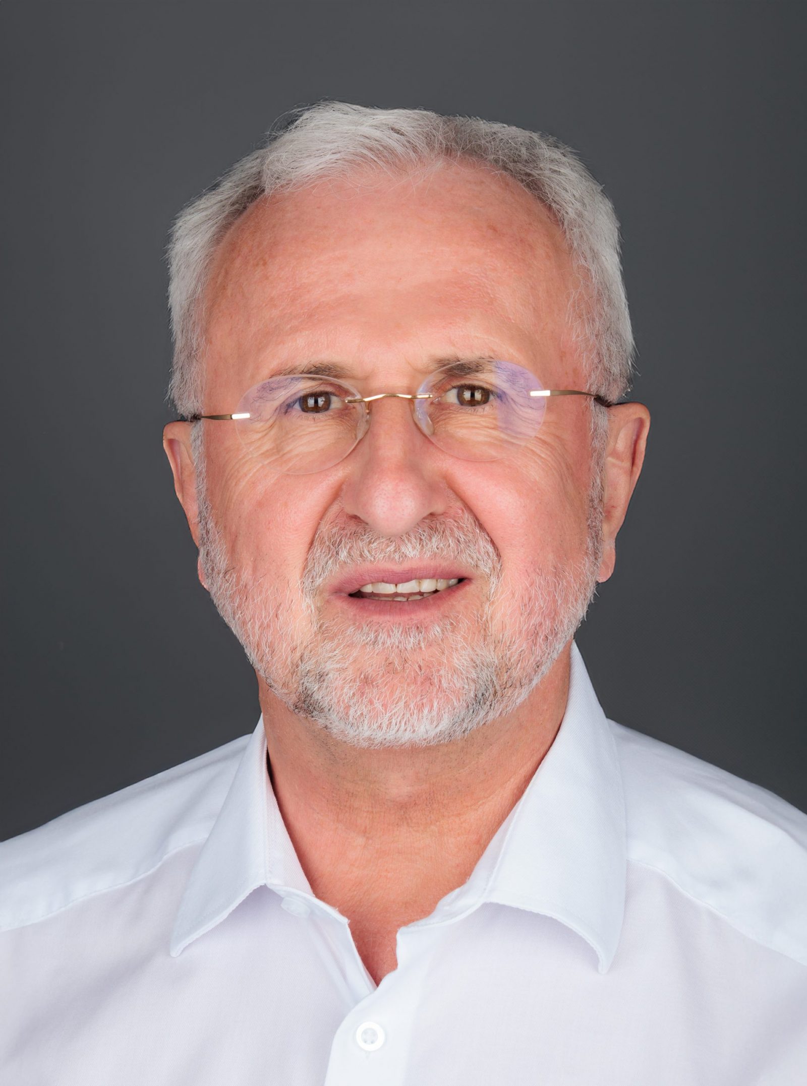 Elderly man with white hair and glasses, wearing a white collared shirt, against a gray background.