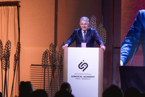 Prof Ninković in suit speaking at a podium labeled "International Surgical Academy Prof. Ninkovic," with decorative leaf designs in the background.