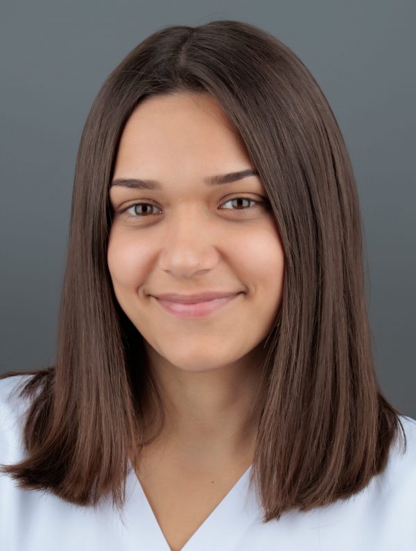 Mlada žena smeđe kose i neutralnog izraza lica, odjevena u bijelu majicu, pozira ispred sive pozadine.