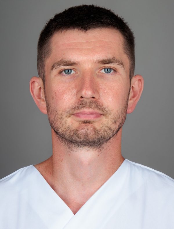 Muškarac kratke tamne kose, svijetloplavih očiju, nosi bijelu majicu, pozira protiv sive pozadine.