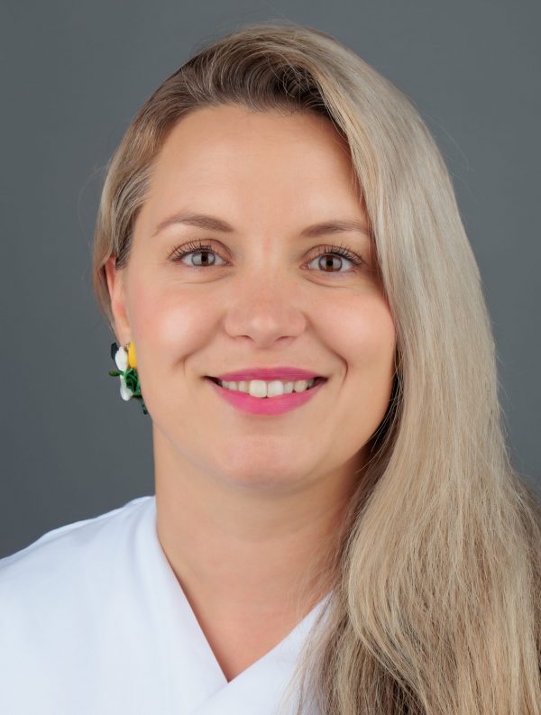 Žena plave kose i zelenih naušnica pozira nasmiješena ispred sive pozadine.