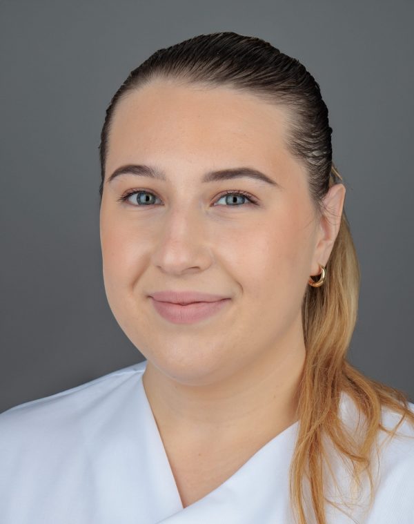 Žena sa svijetlom kosom u bijeloj odjeći, nasmiješena, s naušnicama u obliku obruča, na sivoj pozadini.