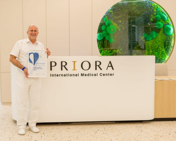 Stariji muškarac u bijeloj odjeći drži priznanje u rukama ispred stola s natpisom 'PRIORA International Medical Center', a u pozadini se vidi zeleni ukrasni detalj na zidu.