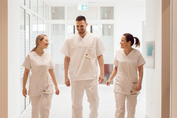 Trojica zdravstvenih radnika u bijelim uniformama nasmiješeni hodaju svijetlim hodnikom bolnice.