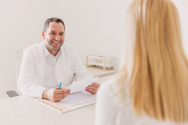 Liječnik u bijelom odijelu sjedi za stolom, držeći papire i razgovarajući sa ženskom pacijenticom čija je plava kosa vidljiva s leđa.