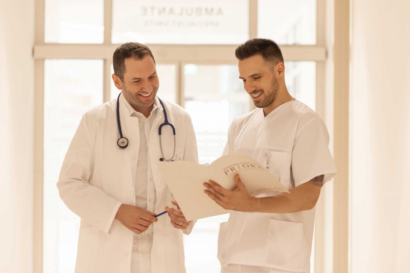 Dva liječnika u hodniku s osmijehom pregledavaju dokumente, jedan drži stetoskop.
