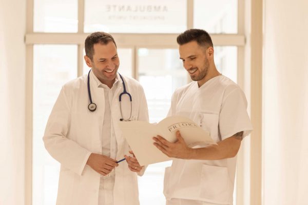Dva liječnika u hodniku s osmijehom pregledavaju dokumente, jedan drži stetoskop.