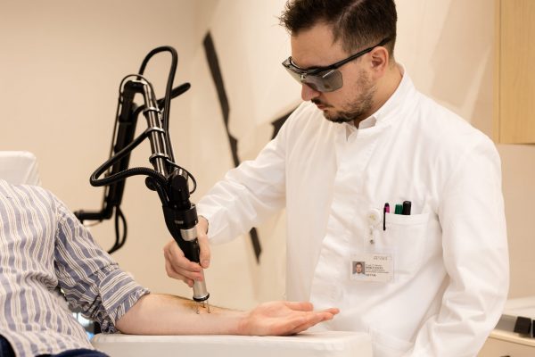Osoba u bijelom mantilu koristi laserski uređaj na ruci druge osobe koja leži na stolu, noseći zaštitne naočale.