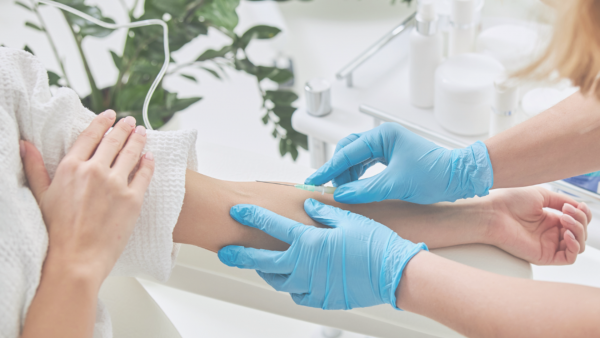 Osoba u plavim rukavicama daje injekciju drugoj osobi na ruci, dok okolina uključuje biljke i bočice.