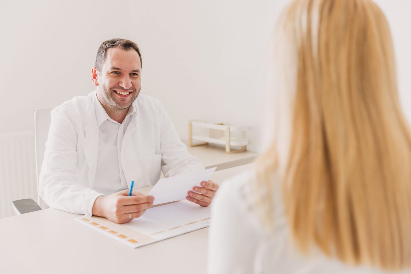 Liječnik u bijelom ogrtaču drži papire i razgovara s pacijenticom u svijetloj prostoriji.