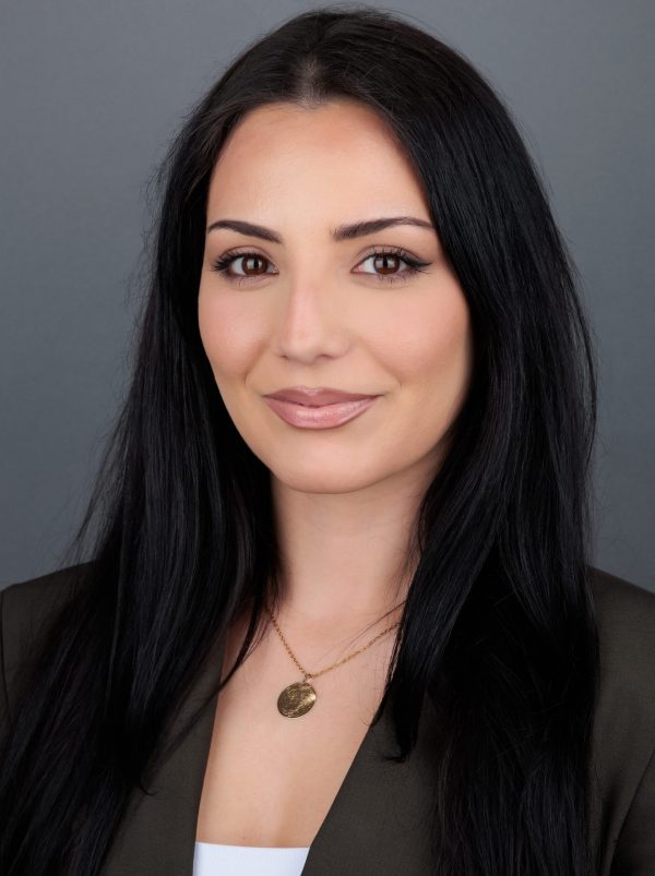 Portret žene s dugom crnom kosom i smeđim očima, koja nosi zlatnu ogrlicu i smeđu jaknu, na neutralnoj sivoj pozadini.