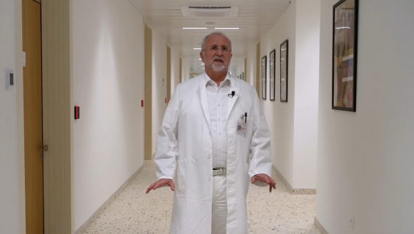 Muškarac u bijelom liječničkom ogrtaču stoji u hodniku, okružen slikama na zidovima.