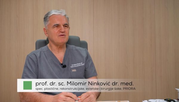 Muškarac u liječničkoj uniformi sjedi i govori ispred drvene pozadine, s natpisom koji navodi njegovo ime i stručnost u plastičnoj, rekonstrukcijskoj, estetskoj kirurgiji ruke.