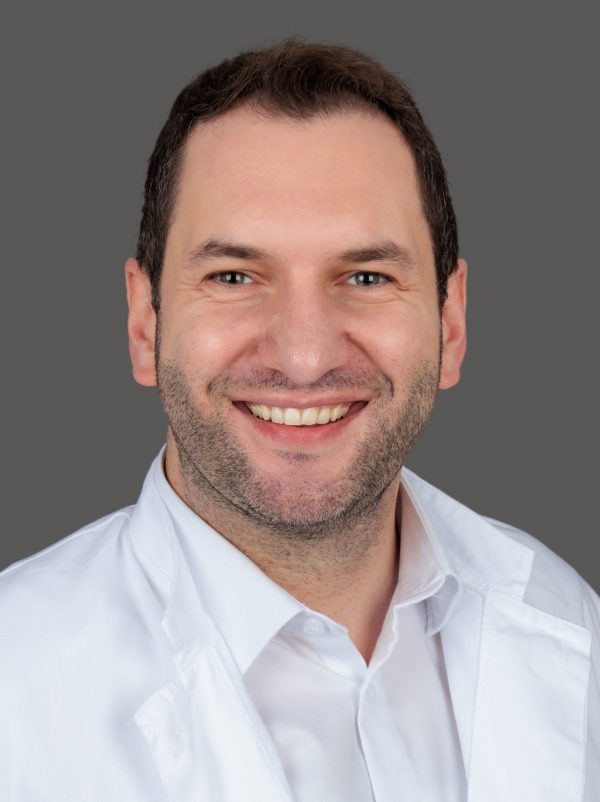 Headshot of a smiling man with short dark hair and light stubble wearing a white coat against a gray background.