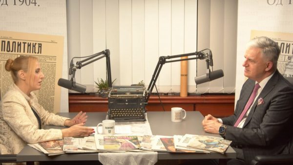 Dvije osobe razgovaraju za stolom s mikrofonima, starinskom pisaćom mašinom, šalicama i novinama, ispred postera s natpisom 'Политика'.