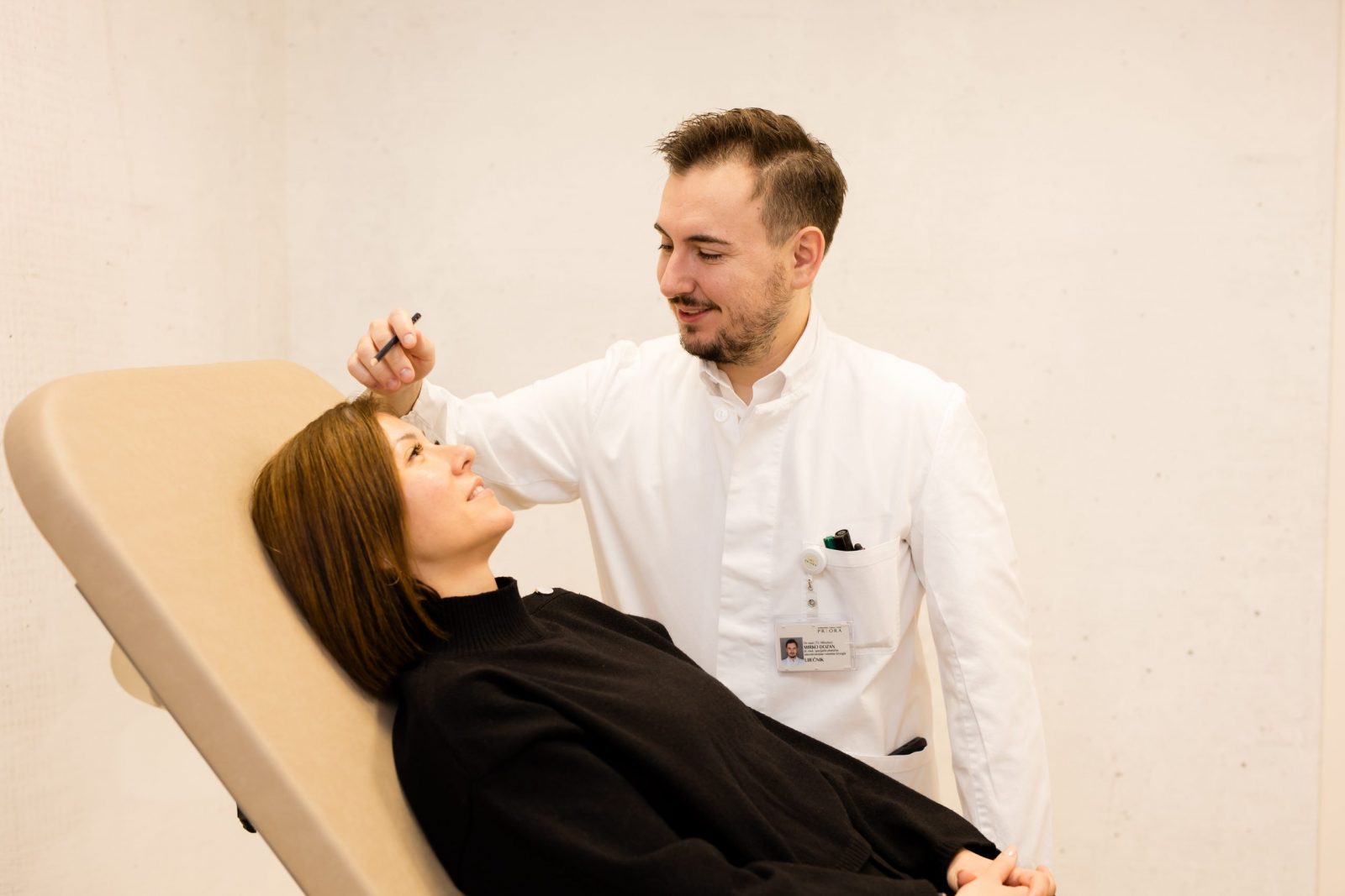 Žena leži na ležaju, dok muškarac u bijeloj liječničkoj odjeći, s oznakom ID kartice na džepu, provodi pregled na njezinom licu držeći mali instrument.