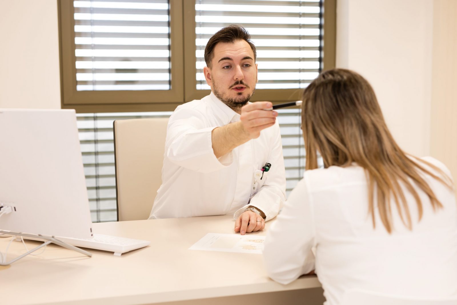 Doctor in a white coat gestures with a pen while speaking to a patient across a desk in a bright office with a computer and papers.