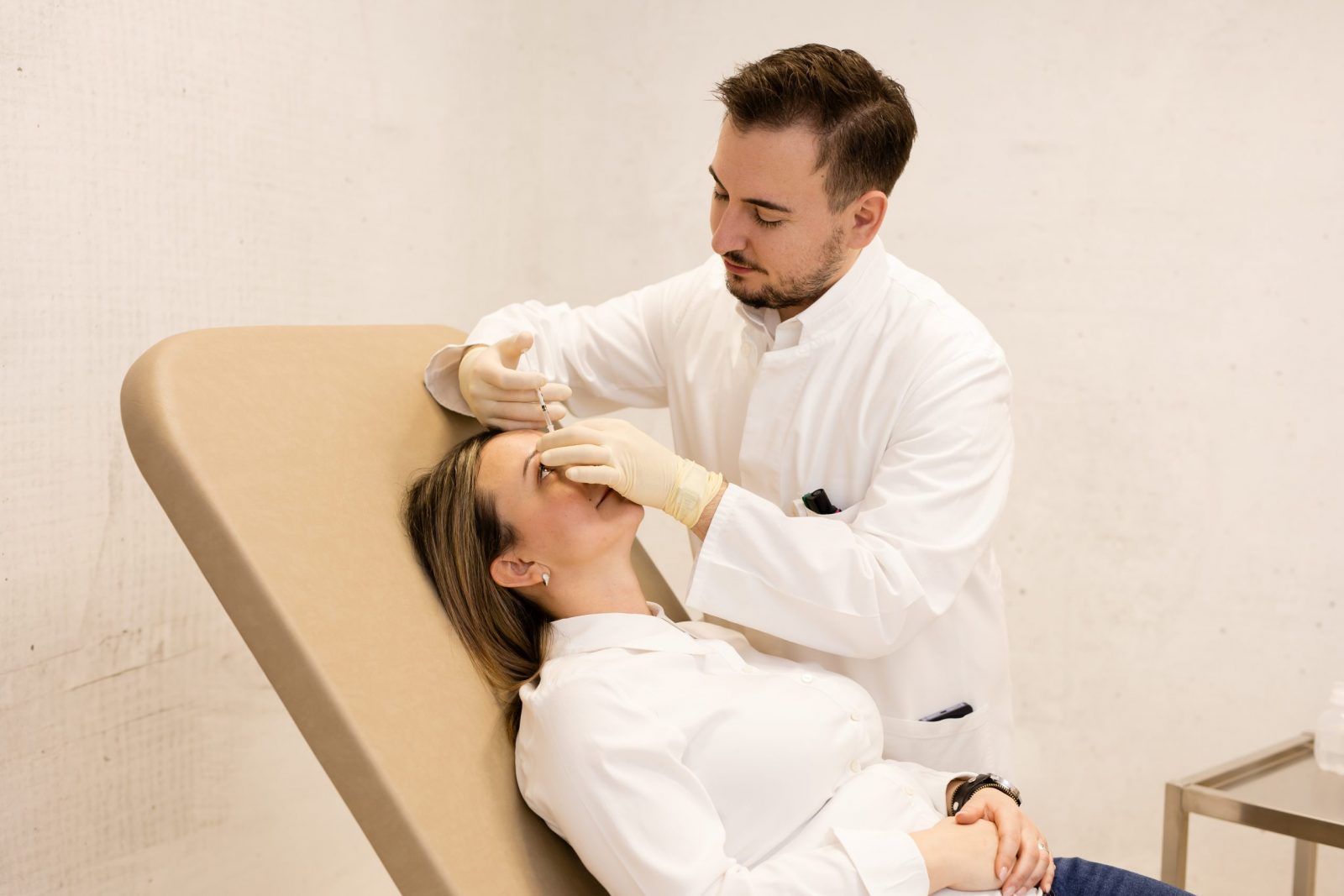 A gloved clinician in a white coat injects a patient’s forehead with a syringe as they recline on an exam chair in a clinic.