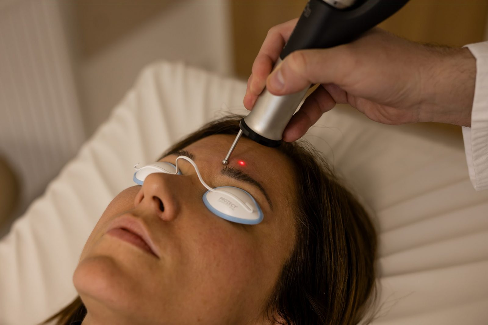 Person wearing protective eye shields receives a laser treatment on the forehead from a handheld device in a clinic.