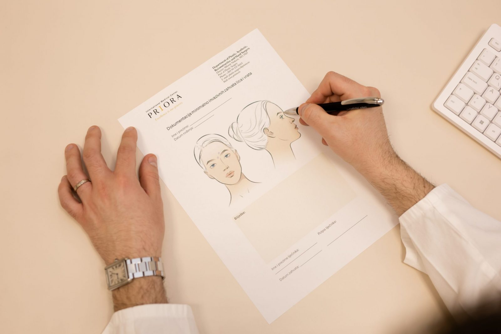 Osoba u bijeloj košulji crta na papiru s ilustracijama ženskog lica sprijeda i sa strane, na stolu s računalnom tipkovnicom u desnom kutu.