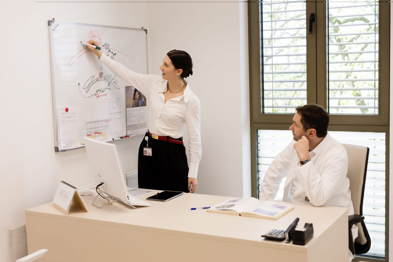Woman pointing at a whiteboard with sketches while a colleague in a white coat listens from behind a desk with a computer, open book, and stationery in a bright office.