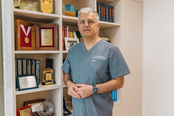 A gray-haired doctor in scrubs stands in an office beside shelves filled with medical books, awards, and plaques.