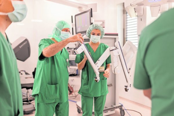 Surgeons in green scrubs calibrate a robotic surgery system in a modern operating room, adjusting articulated arms near a monitor.