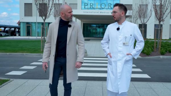 Dva muškarca hodaju preko zebre ispred zgrade s natpisom 'International Medical Center'. Lijevi muškarac nosi kaput svijetle boje, a desni nosi bijeli liječnički mantil.