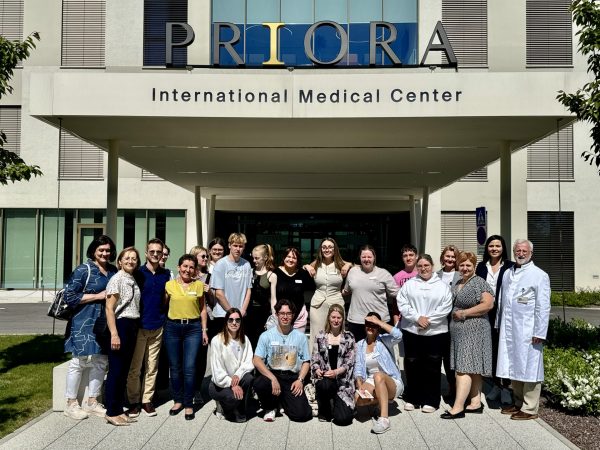 Large group of people pose under the entrance canopy of a building with signage reading “PRIORA International Medical Center” on a sunny day, including one person in a white lab coat.