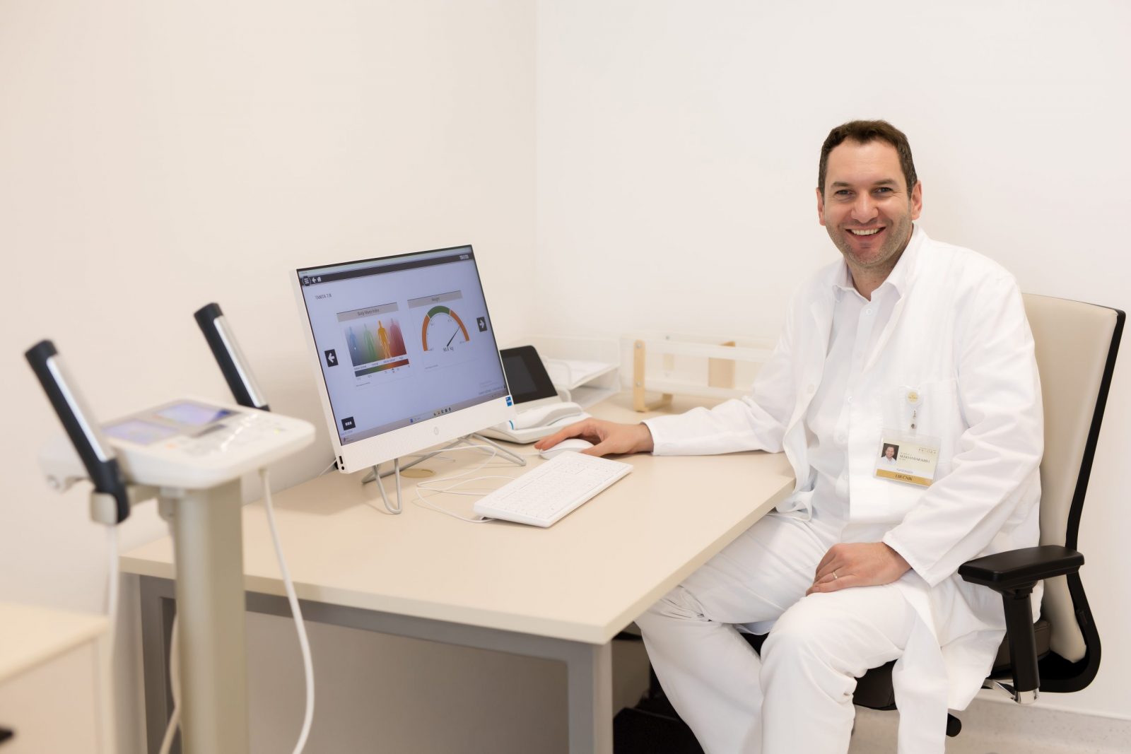 Smiling doctor in a white coat sits at a desk in a bright clinic office, with a computer displaying health metrics charts and a nearby medical device.