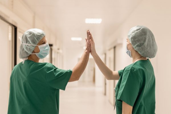 Dvije medicinske sestre u zelenim uniformama i zaštitnim maskama na licu, daju si high-five u bolničkom hodniku.