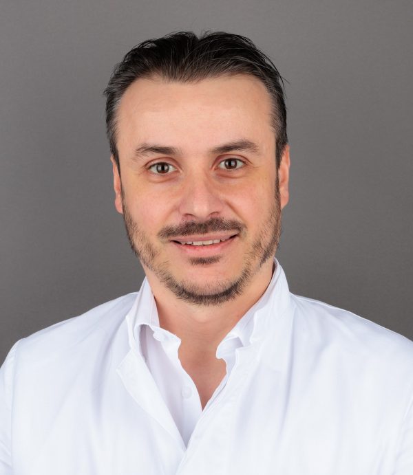 Muškarac s kratkom crnom kosom i bradom, odjeven u bijelu košulju, smiješi se ispred sive pozadine.