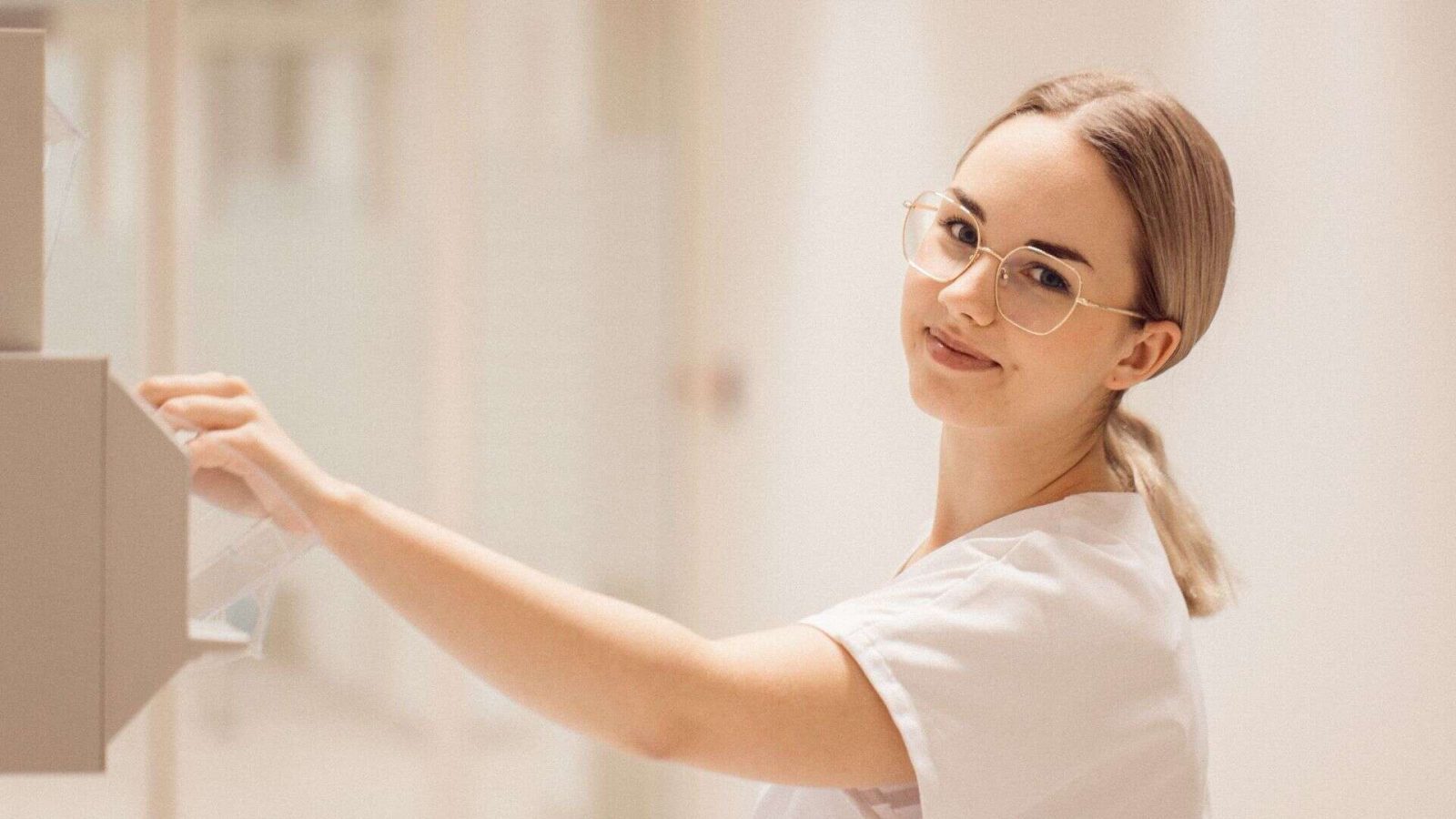 Smiling healthcare worker in white scrubs and glasses reaches for supplies from a wall dispenser in a bright hospital corridor.