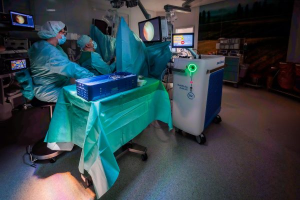 Two surgeons in blue sterile gowns and masks perform a minimally invasive procedure on a draped patient in a dimly lit operating room, guided by endoscopic monitors with a laser system beside the table.