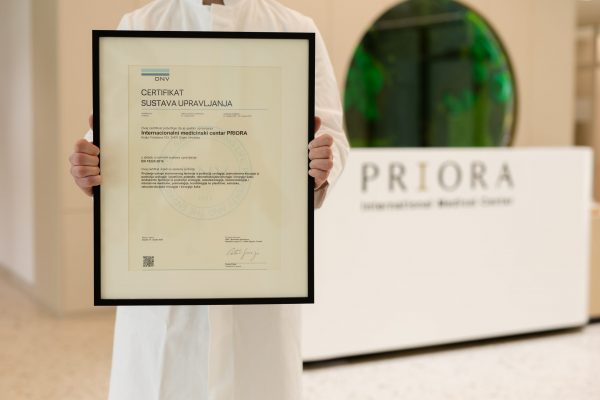 Person in a white coat holds a framed DNV management system certificate for the International Medical Center PRlORA, with the PRlORA reception desk visible in the clinic lobby background.