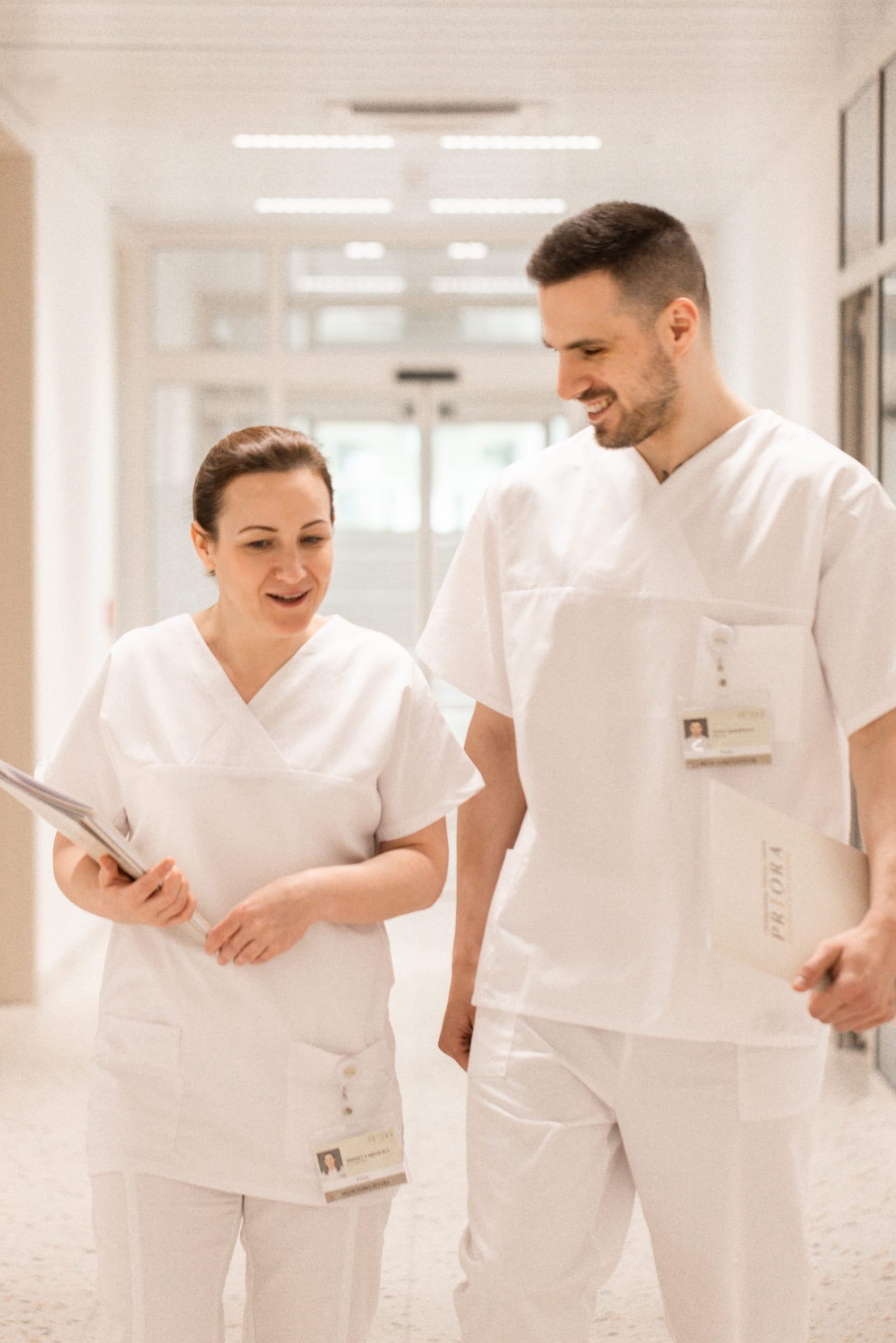 Two healthcare workers in white scrubs walk down a bright hospital hallway, smiling and talking while carrying folders and wearing ID badges.