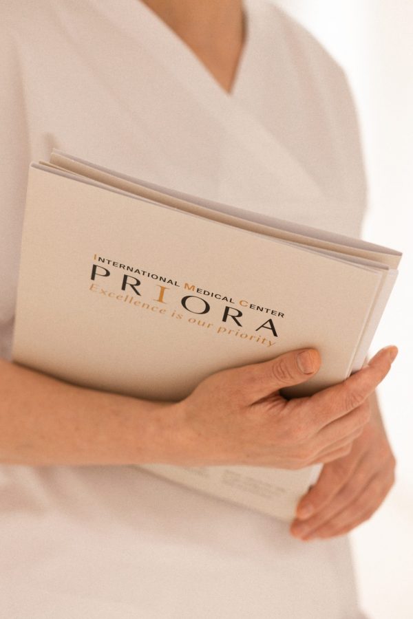 Osoba u bijeloj odjeći drži dokument s logotipom 'International Medical Center PRIORA' i sloganom 'Excellence is our priority'.