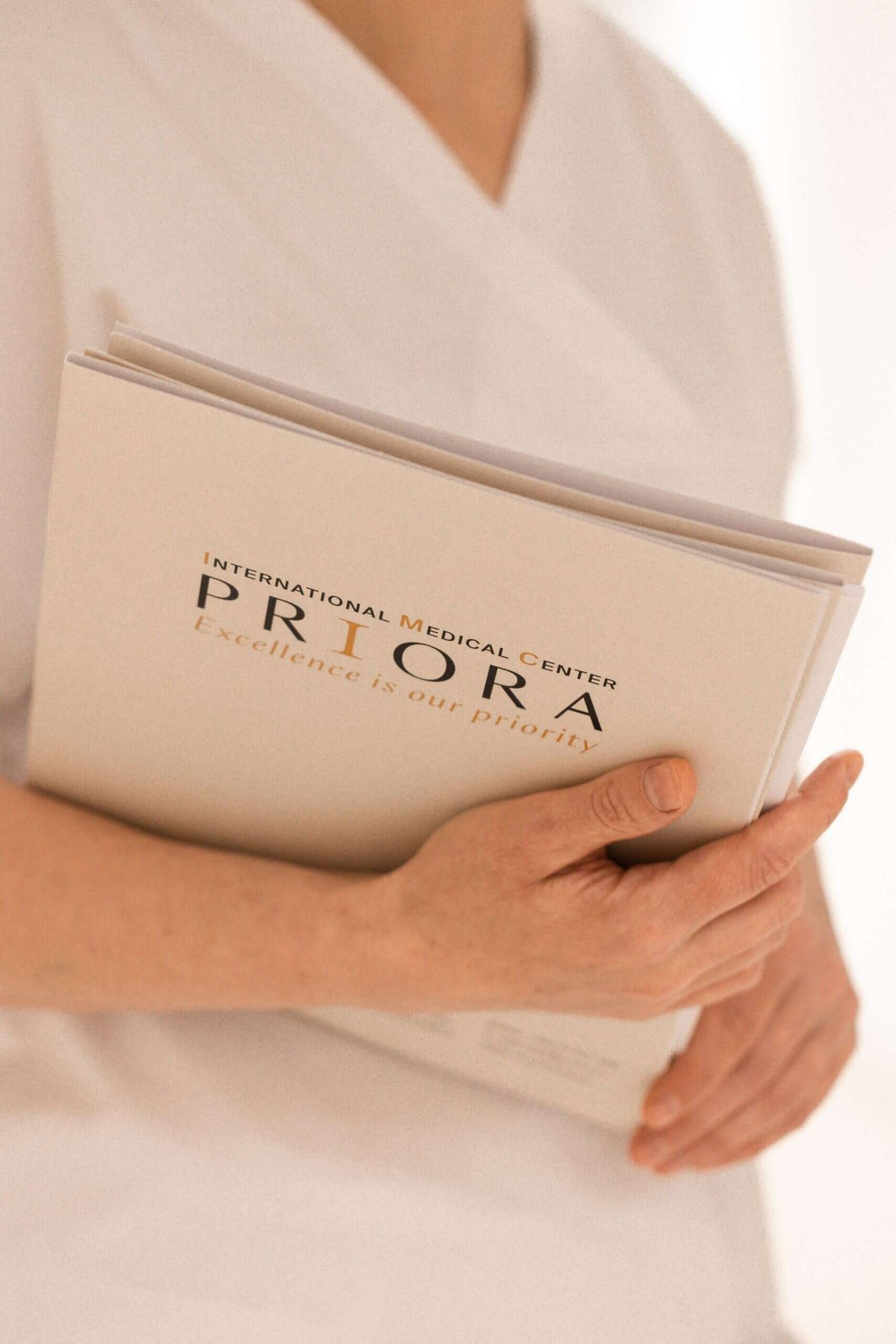 A person in a white uniform holds a beige brochure reading “International Medical Center PRIORA – Excellence is our priority” against a soft, minimal background.