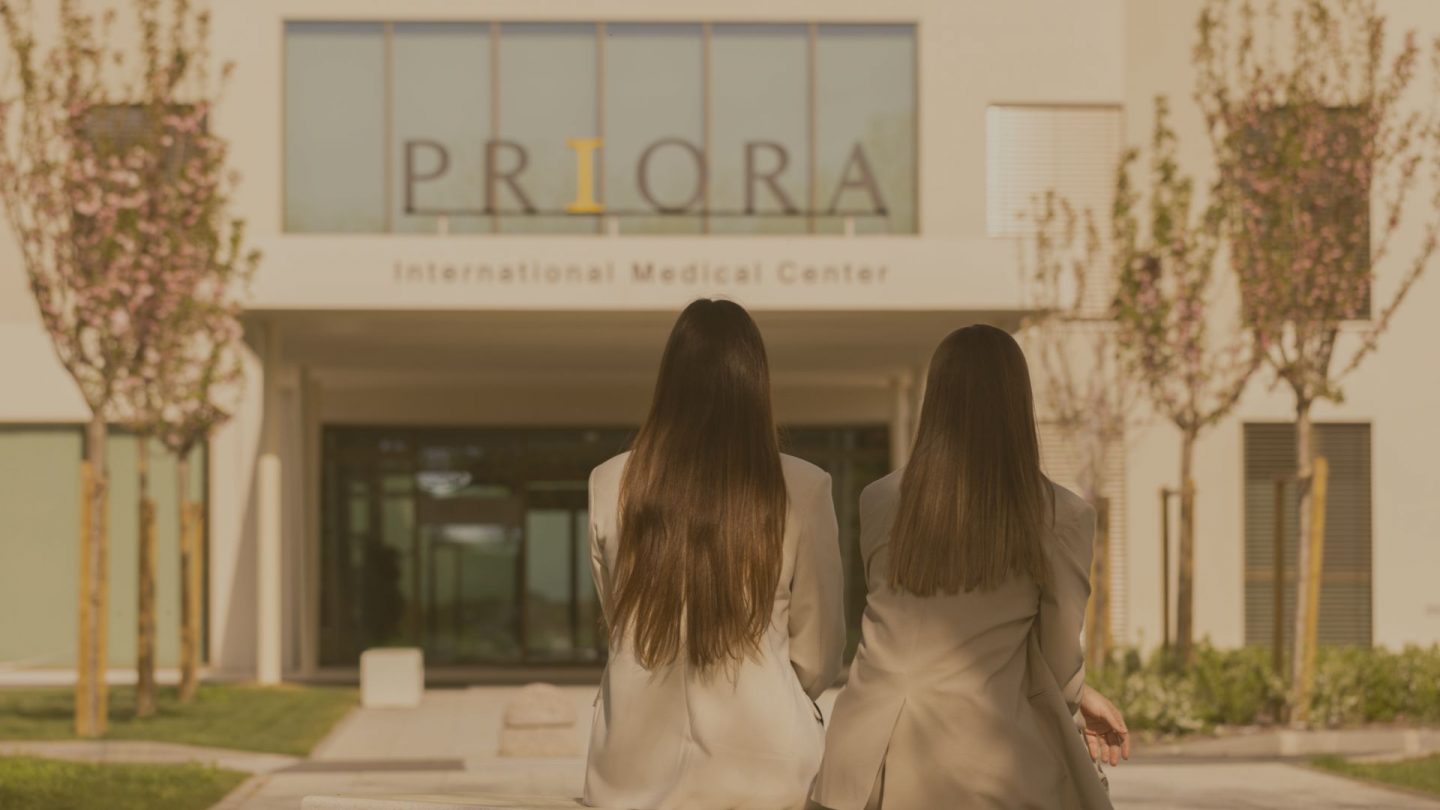 Dvije žene s dugom tamnom kosom sjede na klupi ispred ulaza u zgradu s natpisom 'PRIORA International Medical Center' na fasadi.