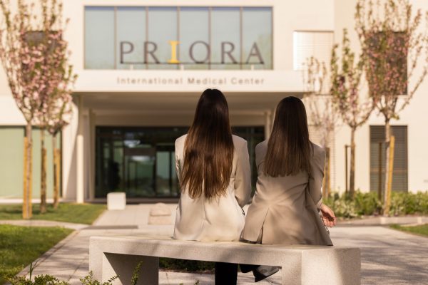 Dva čovjeka s dugom smeđom kosom sjede na kamenu klupu ispred međunarodnog medicinskog centra s natpisom 'PRIORA'.