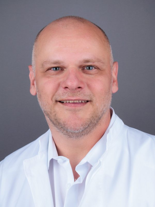 Muškarac obrijane glave u bijeloj liječničkoj kuti na sivom pozadinskom platnu.