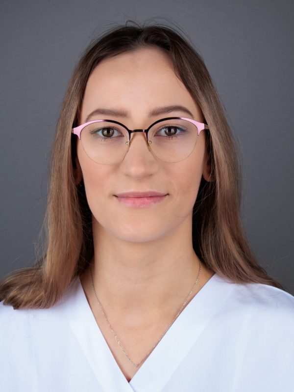 Žena s dugom smeđom kosom nosi naočale i bijelu košulju, protiv tamne pozadine.